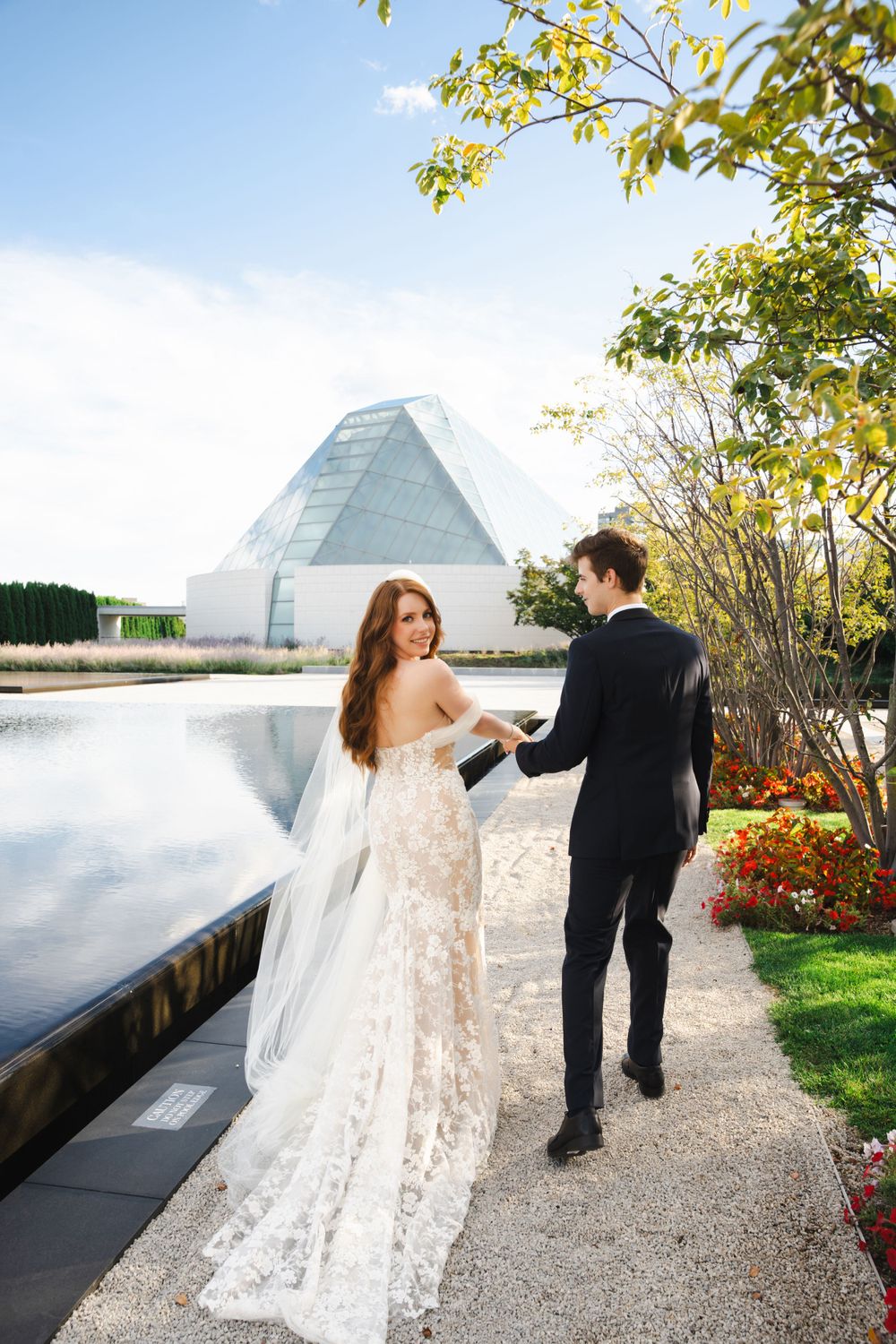 A couple shares a romantic moment by a modern glass pyramid building on their wedding day.