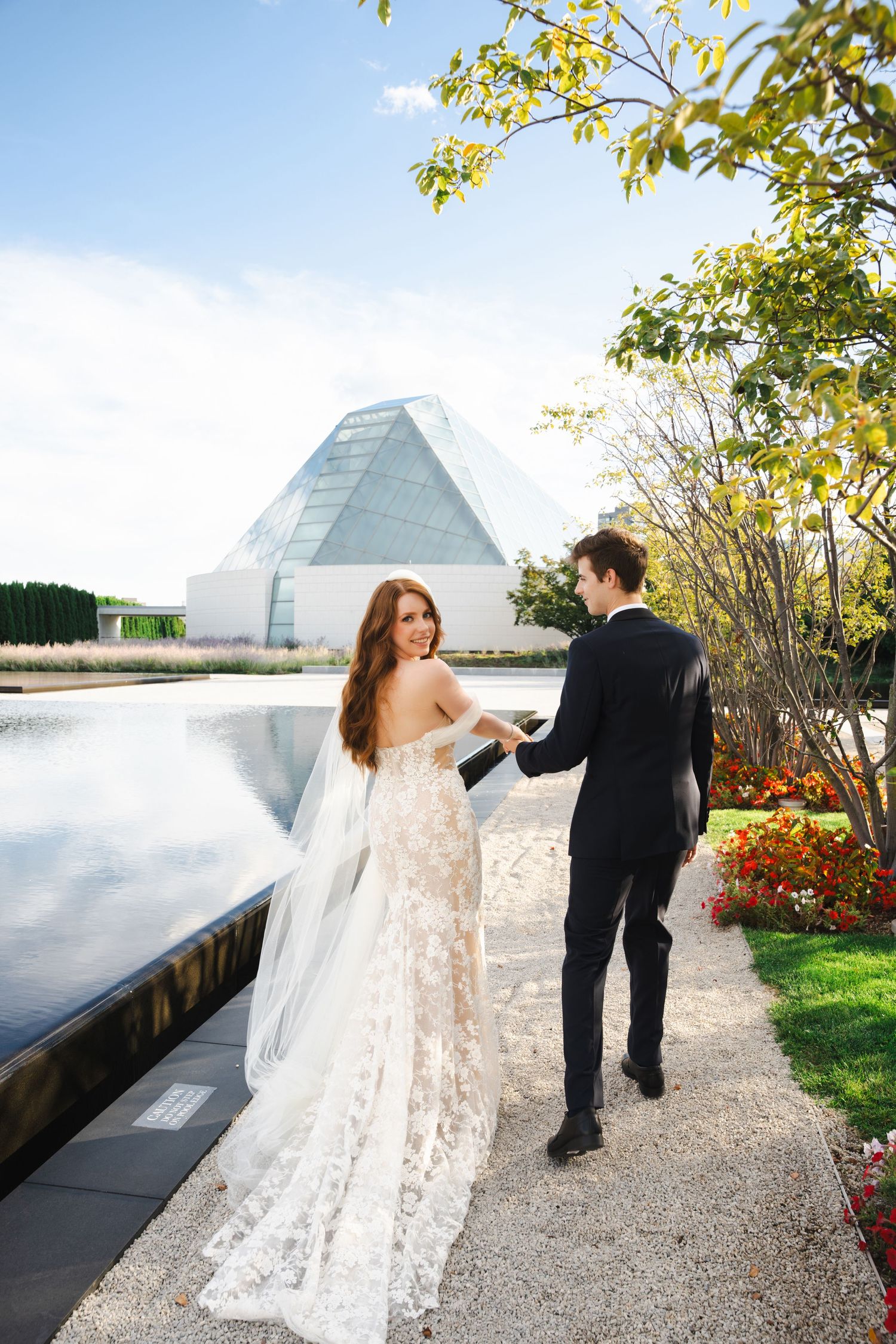 A couple shares a romantic moment by a modern glass pyramid building on their wedding day.