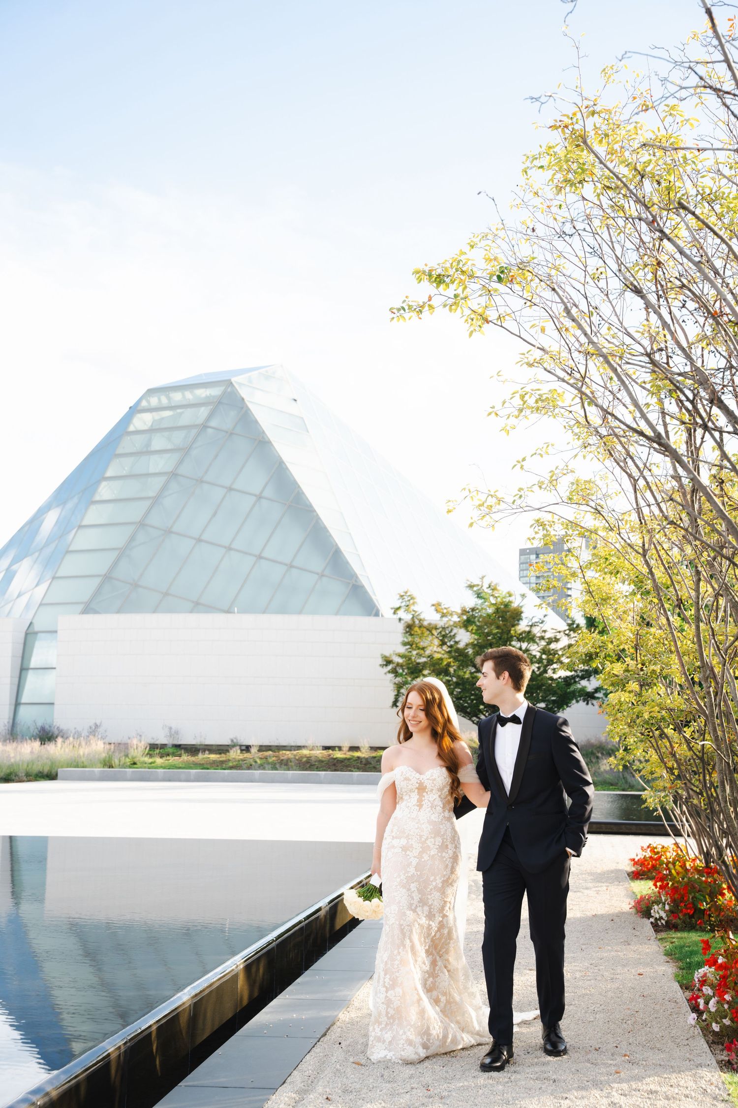 Newlyweds walk along a reflective pool beside a contemporary glass pyramid structure.