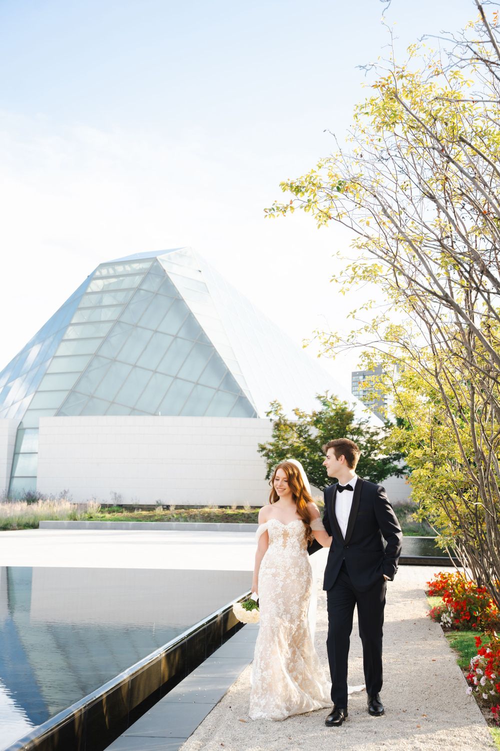 Newlyweds walk along a reflective pool beside a contemporary glass pyramid structure.
