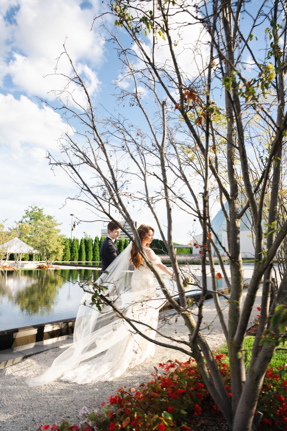 Wedding couple poses beneath autumn trees near a reflective pool and modern architecture.