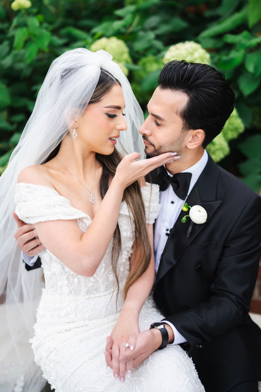 A romantic wedding moment captured between a bride in white lace and groom in black tuxedo against a green garden backdrop.