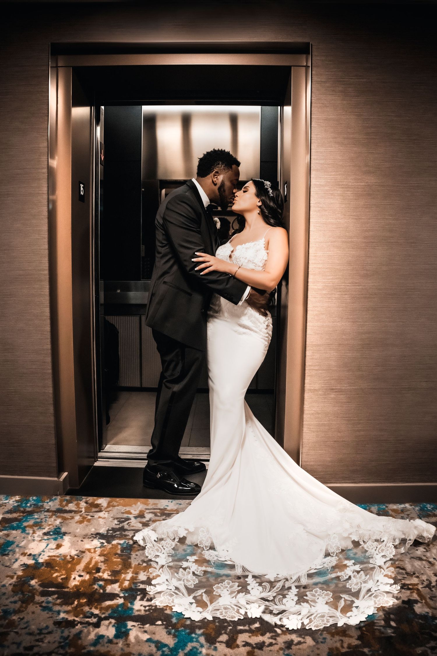 Romantic couple embrace in elevator wearing wedding attire with trailing dress.