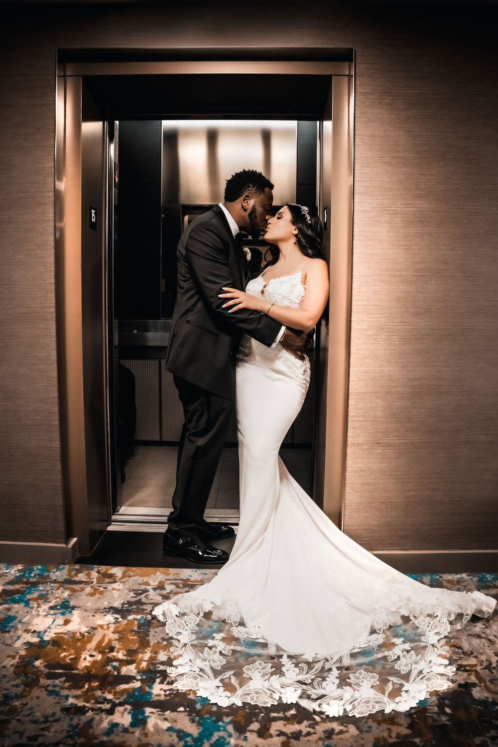 Romantic couple embrace in elevator wearing wedding attire with trailing dress.