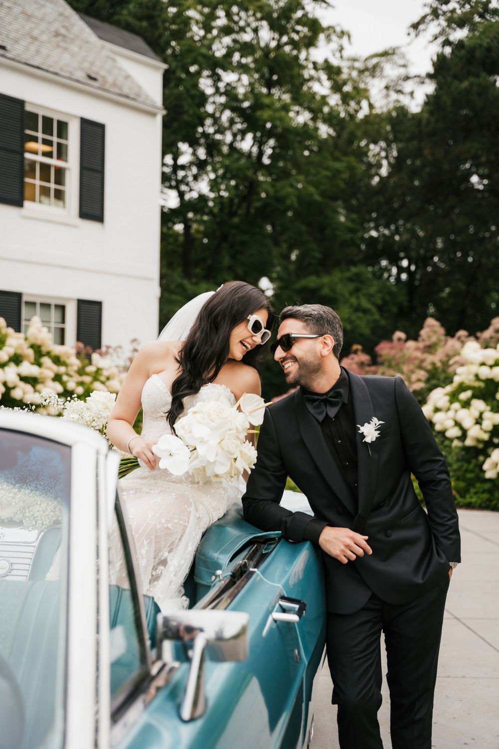 Romantic wedding couple poses with vintage blue car in elegant outdoor setting.