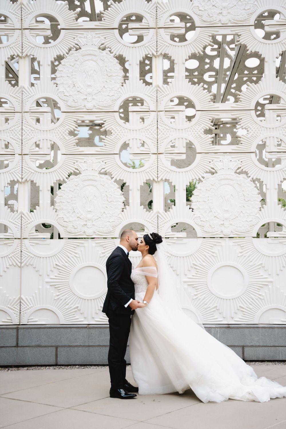 Wedding couple shares intimate moment against ornate white architectural wall.