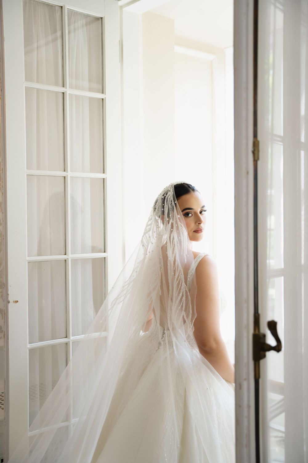 Bride in white veil poses elegantly by French doors in natural light.