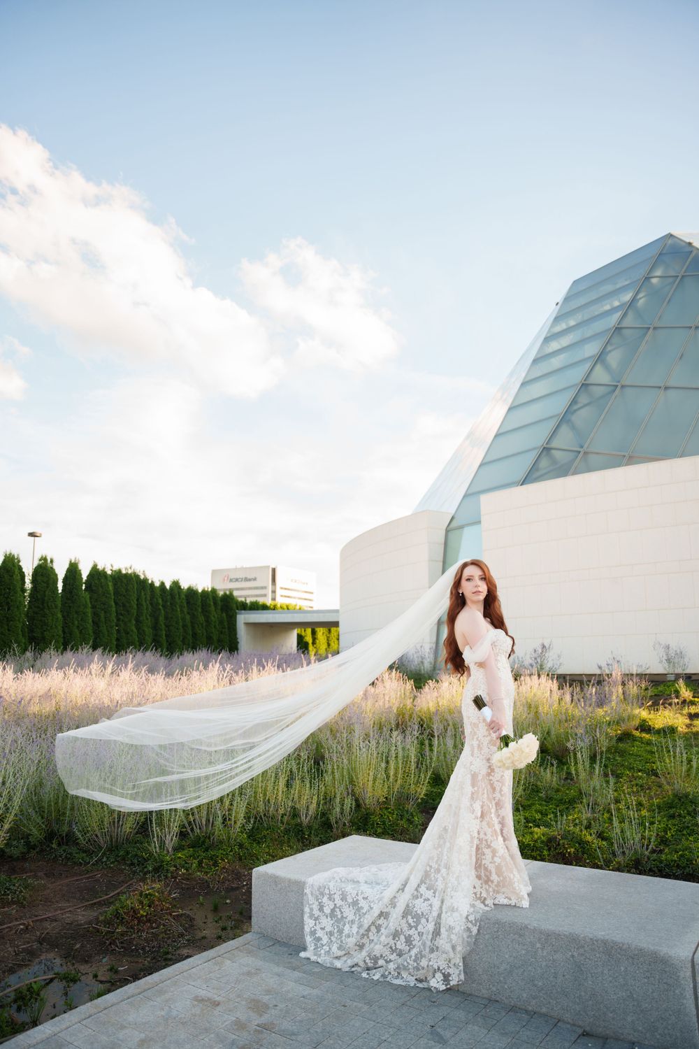 Bride in lace gown with flowing veil poses against modern glass building architecture.