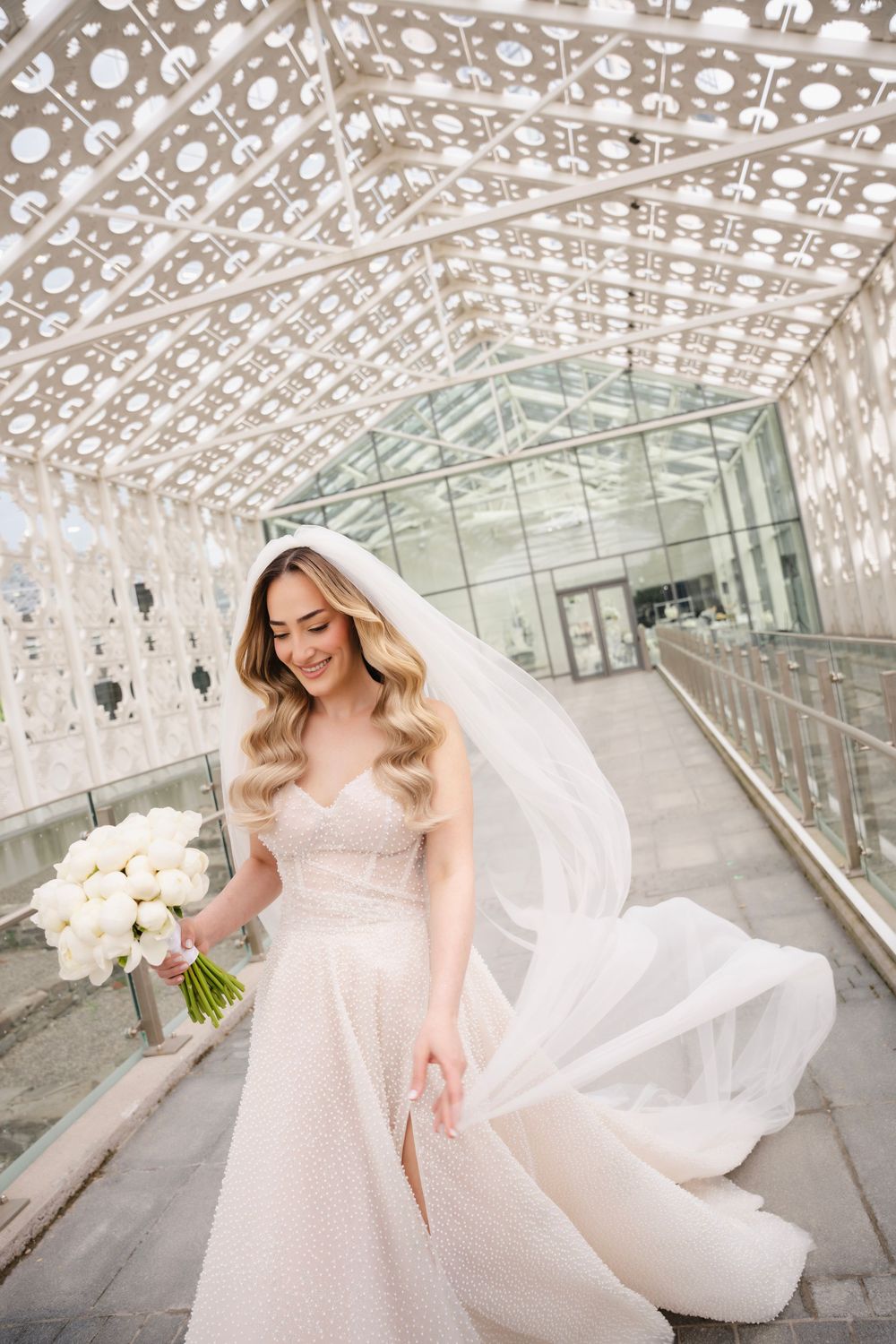 Bride in flowing white gown holding bouquet under geometric patterned architectural structure.