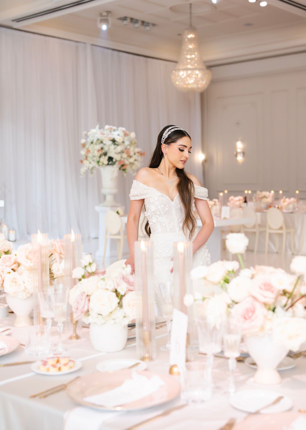 Bride in off-shoulder gown stands amid luxurious white and pink wedding reception decor.