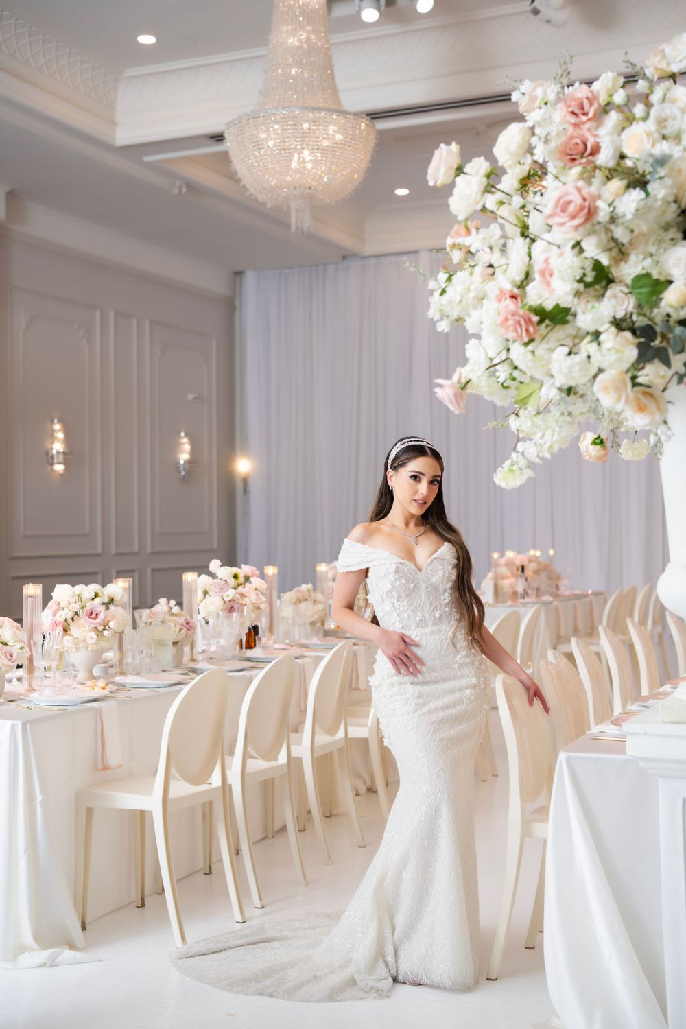 Bride in fitted white gown stands in elegant all-white reception room with crystal chandeliers.