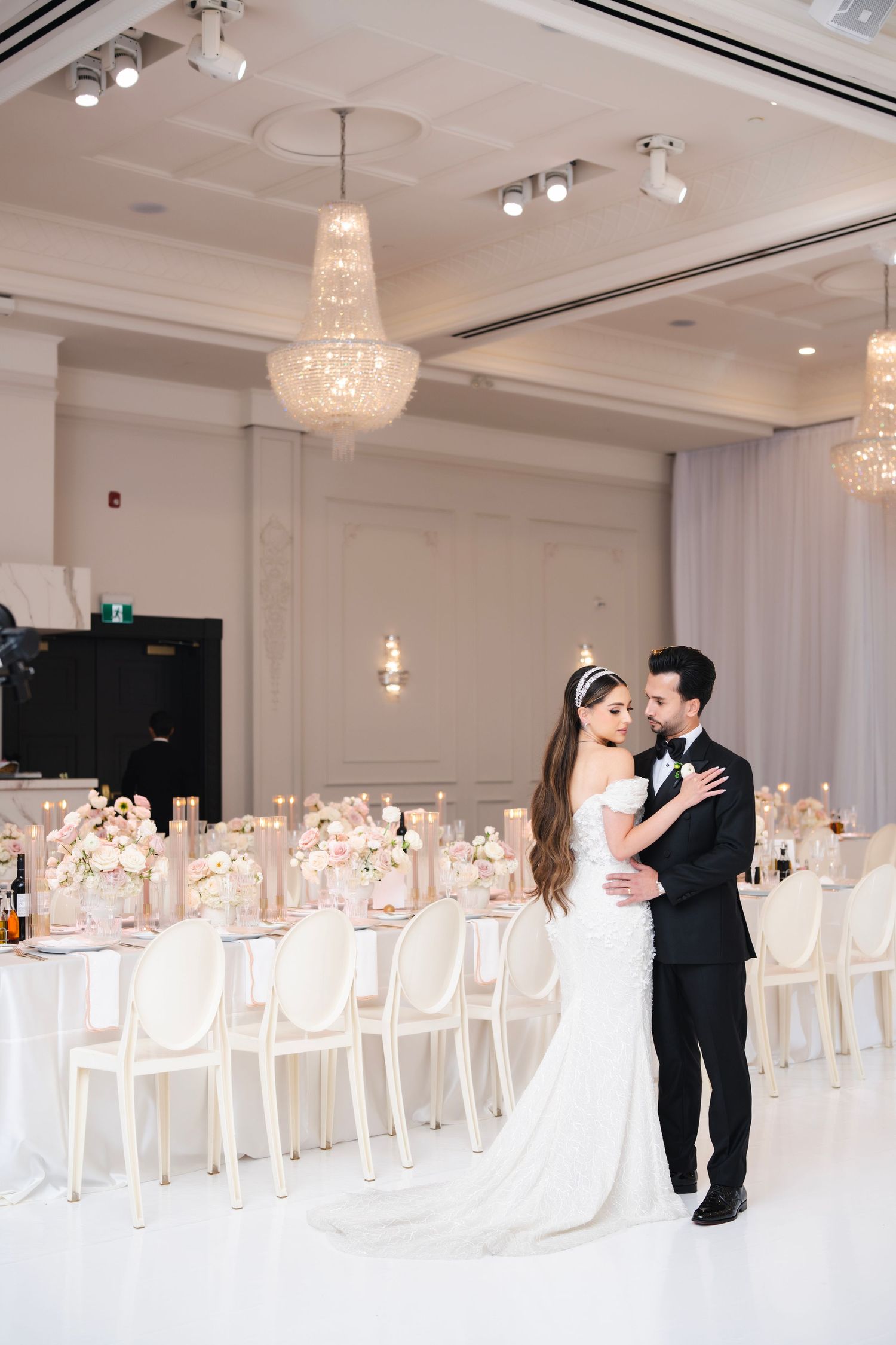 Wedding couple shares intimate dance in elegant white reception hall with crystal chandeliers.