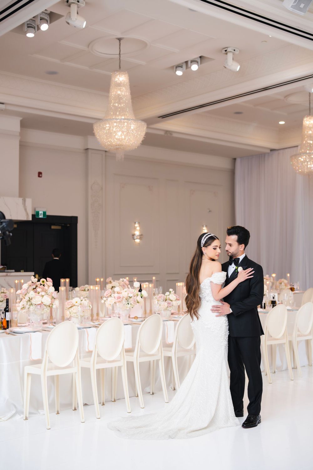 Wedding couple shares intimate dance in elegant white reception hall with crystal chandeliers.