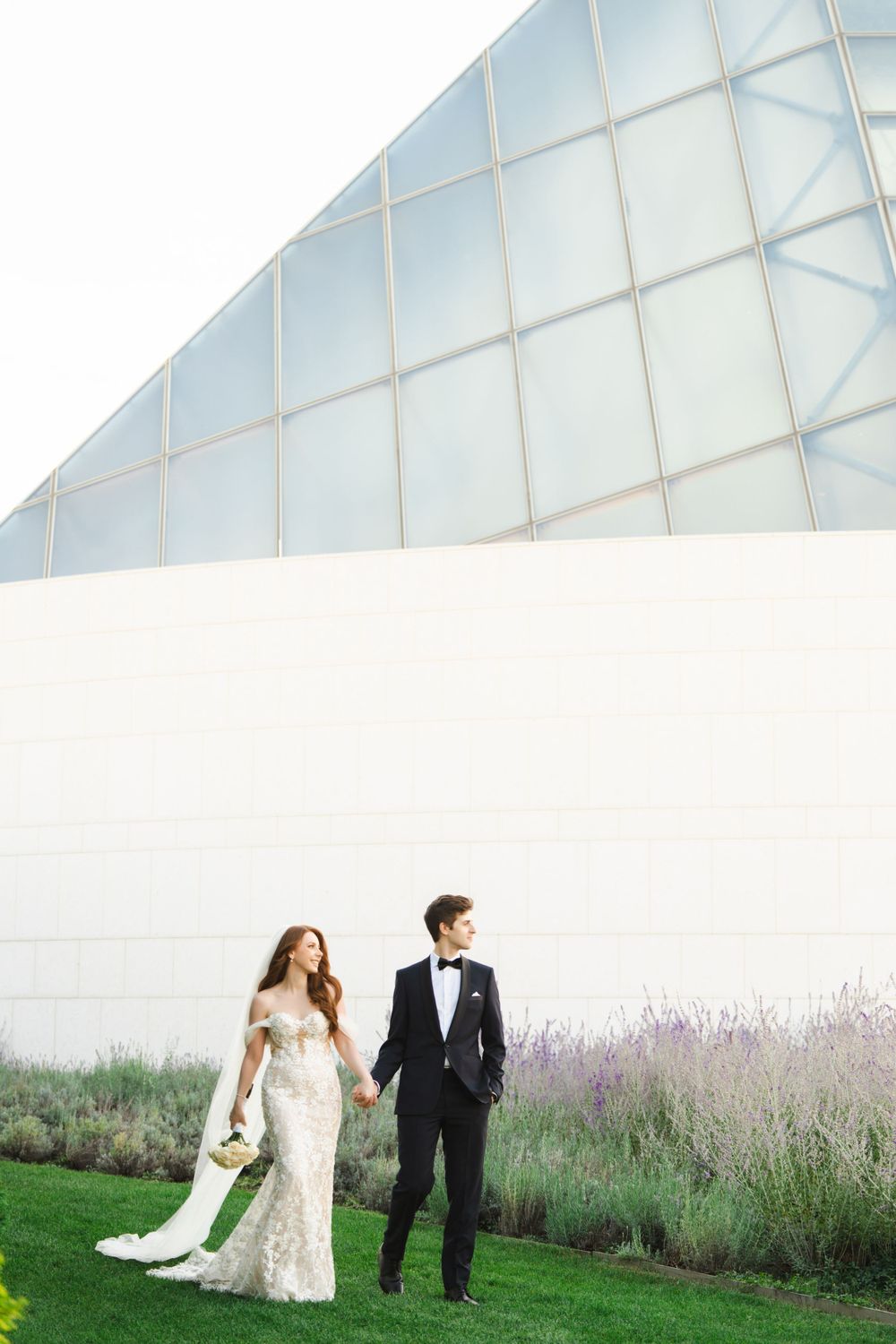 Bride and groom walking beside geometric white building with landscaped garden.