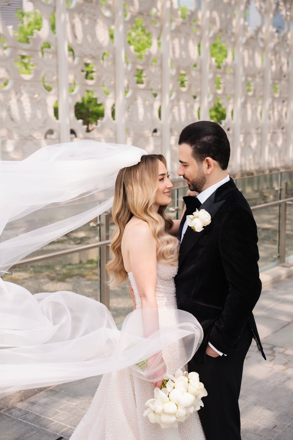 Romantic wedding portrait with bride's veil flowing in breeze under modern architectural setting.