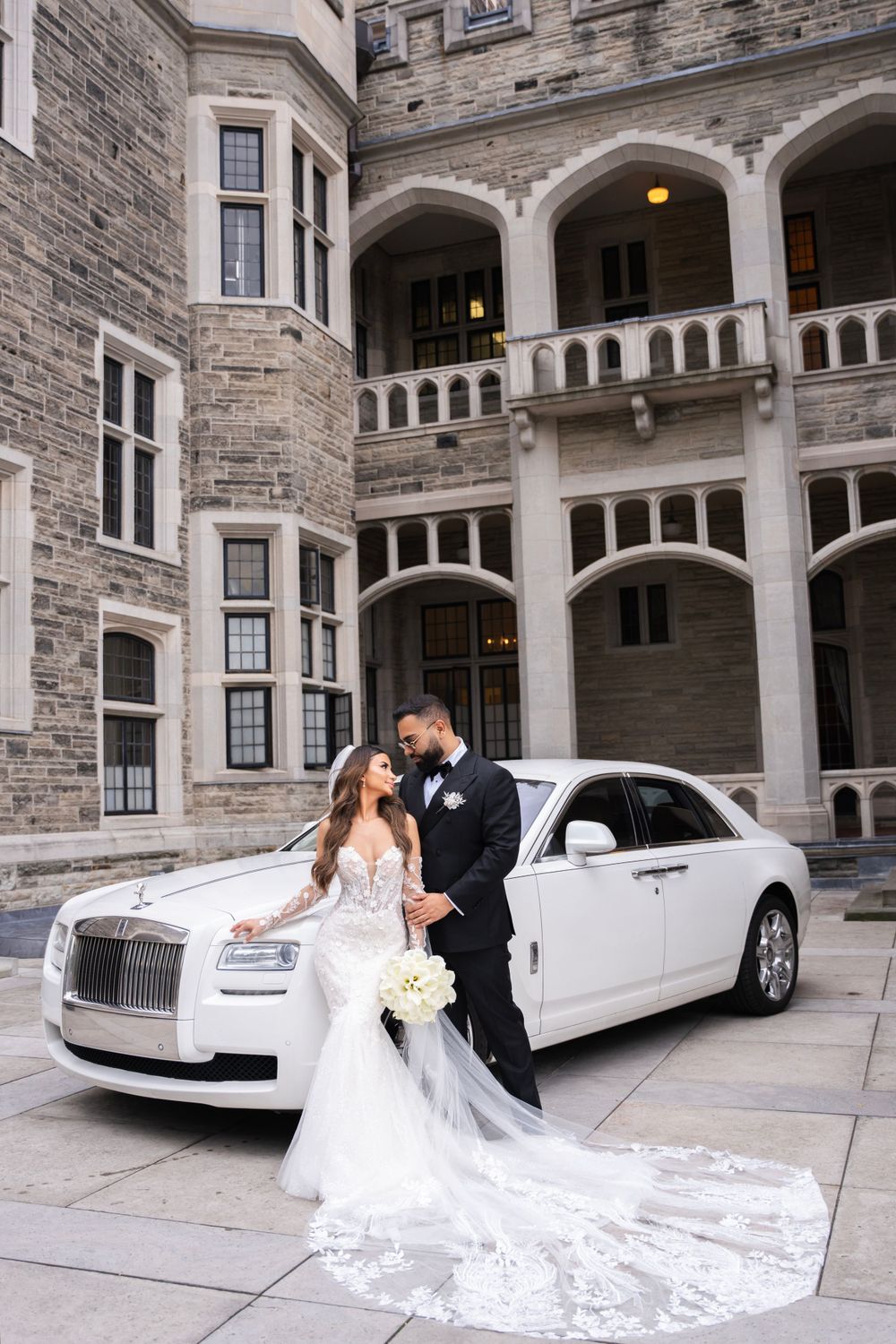 Wedding couple posing with white Rolls Royce outside stone castle building.