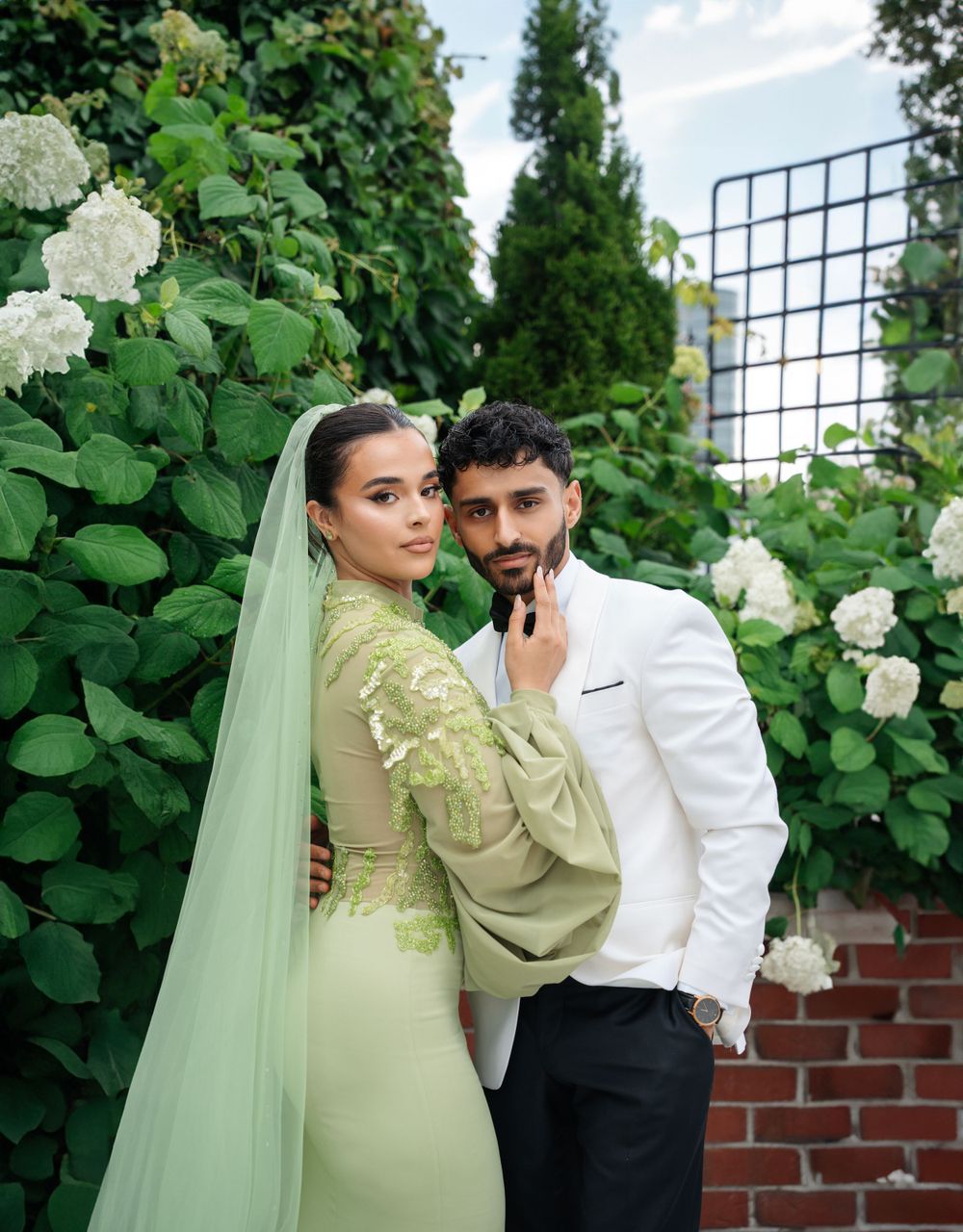 Wedding couple poses intimately against a garden wall with white hydrangeas and greenery.