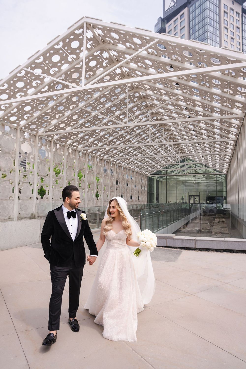 Newlyweds walking under modern geometric architectural canopy in formal wedding attire.