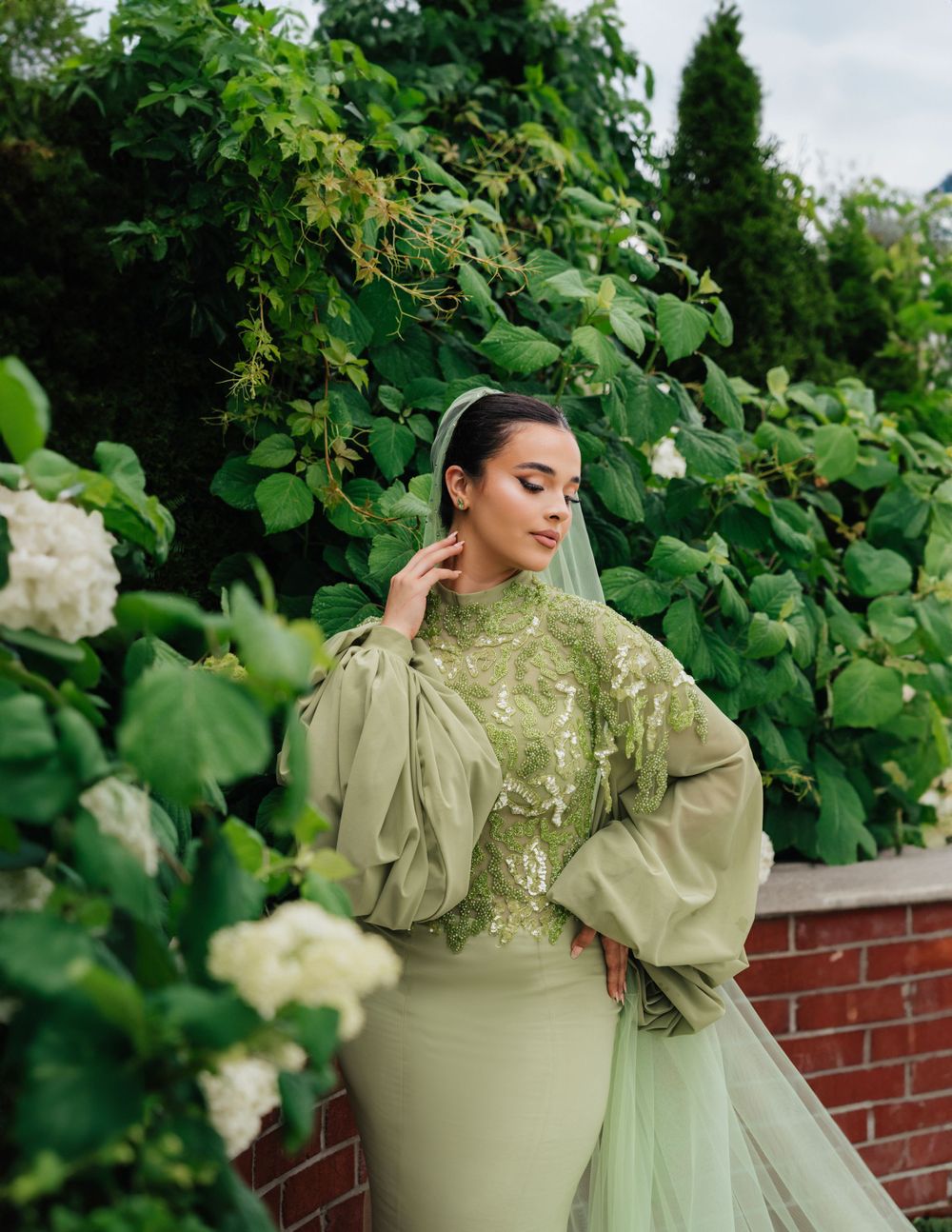 Bride in mint green dress poses gracefully in a lush garden setting with white flowers.