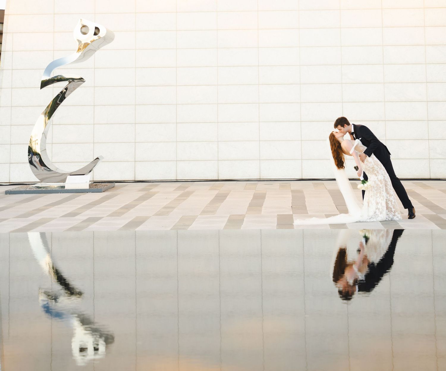 Wedding couple reflected in mirrored surface against white architectural wall.