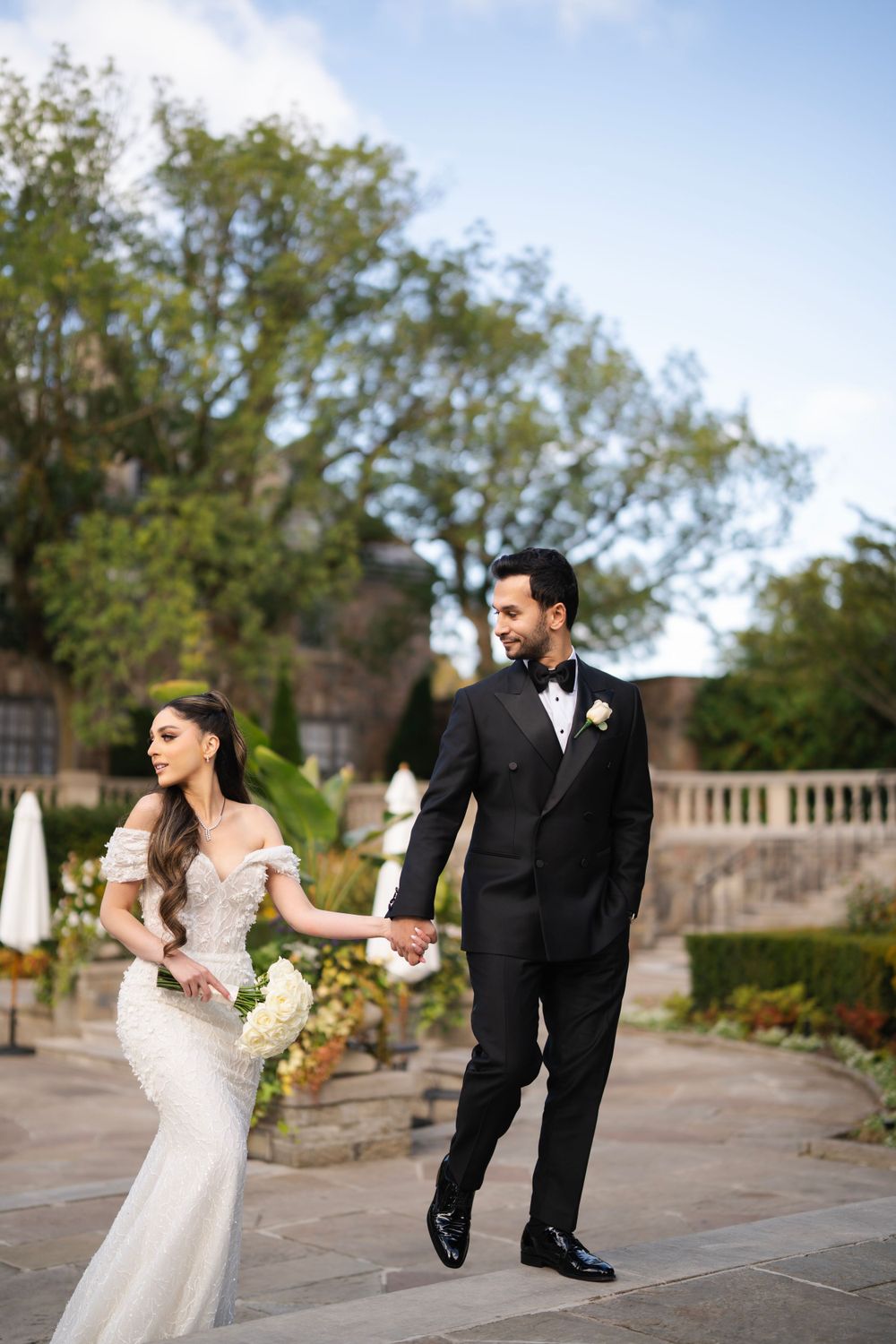 A couple in formal wedding attire walks along a stone terrace during golden hour.