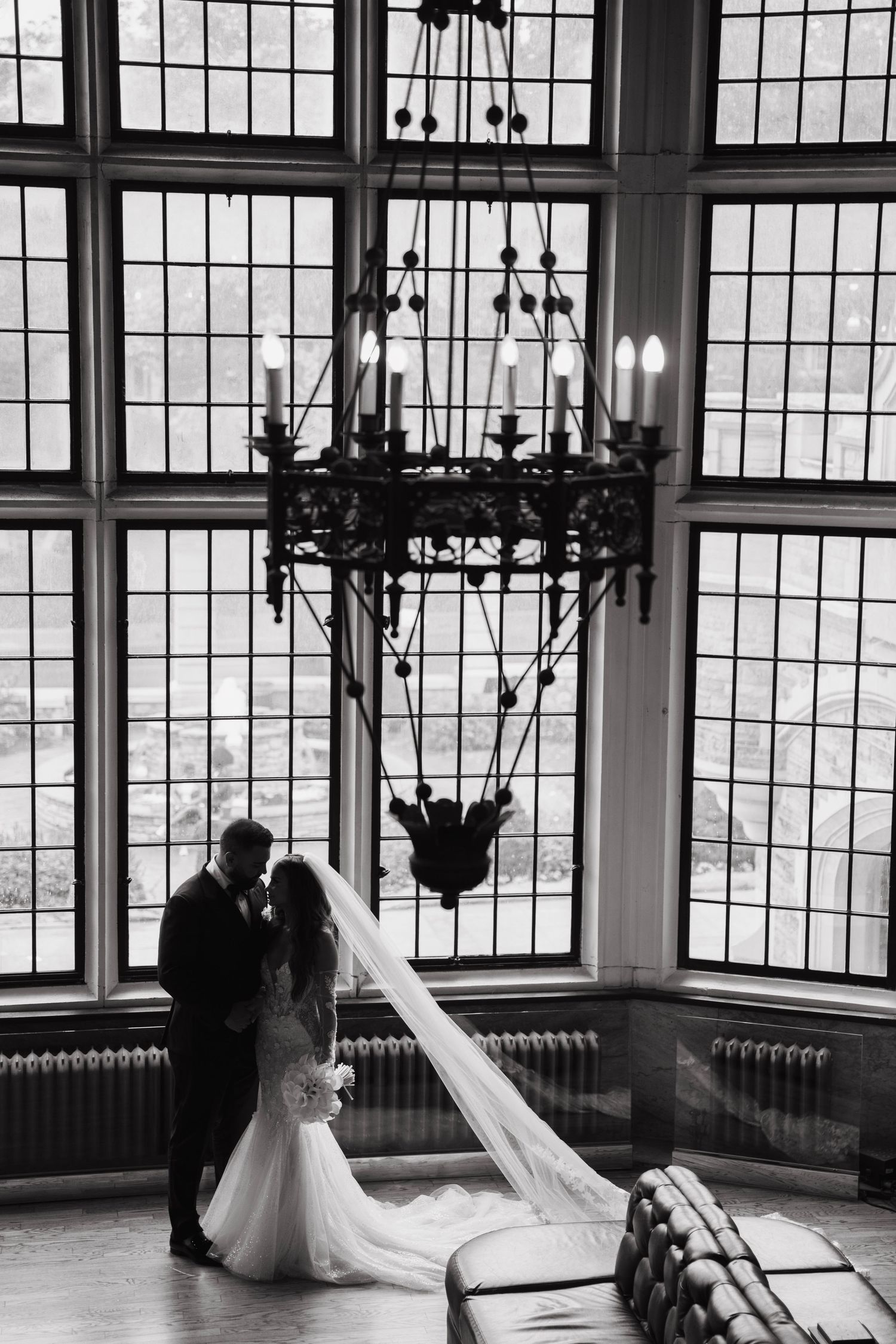 Silhouetted wedding couple standing by large windows with dramatic chandelier.