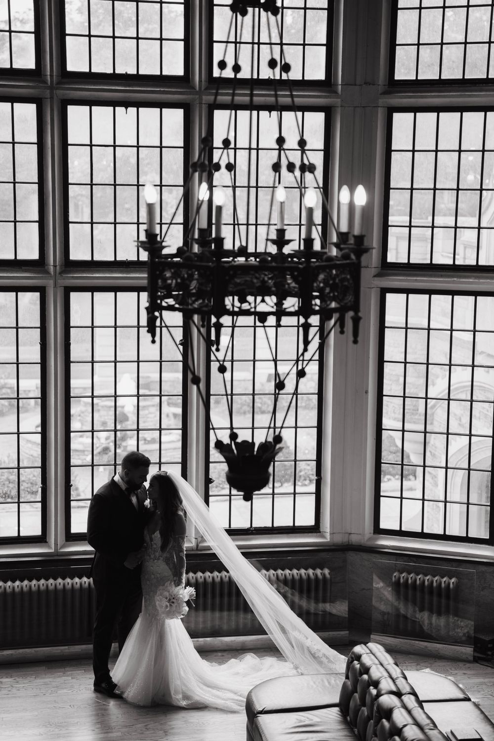 Silhouetted wedding couple standing by large windows with dramatic chandelier.