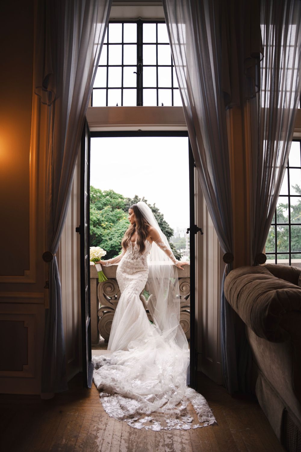 Bride in fitted lace gown silhouetted in window frame with flowing veil.