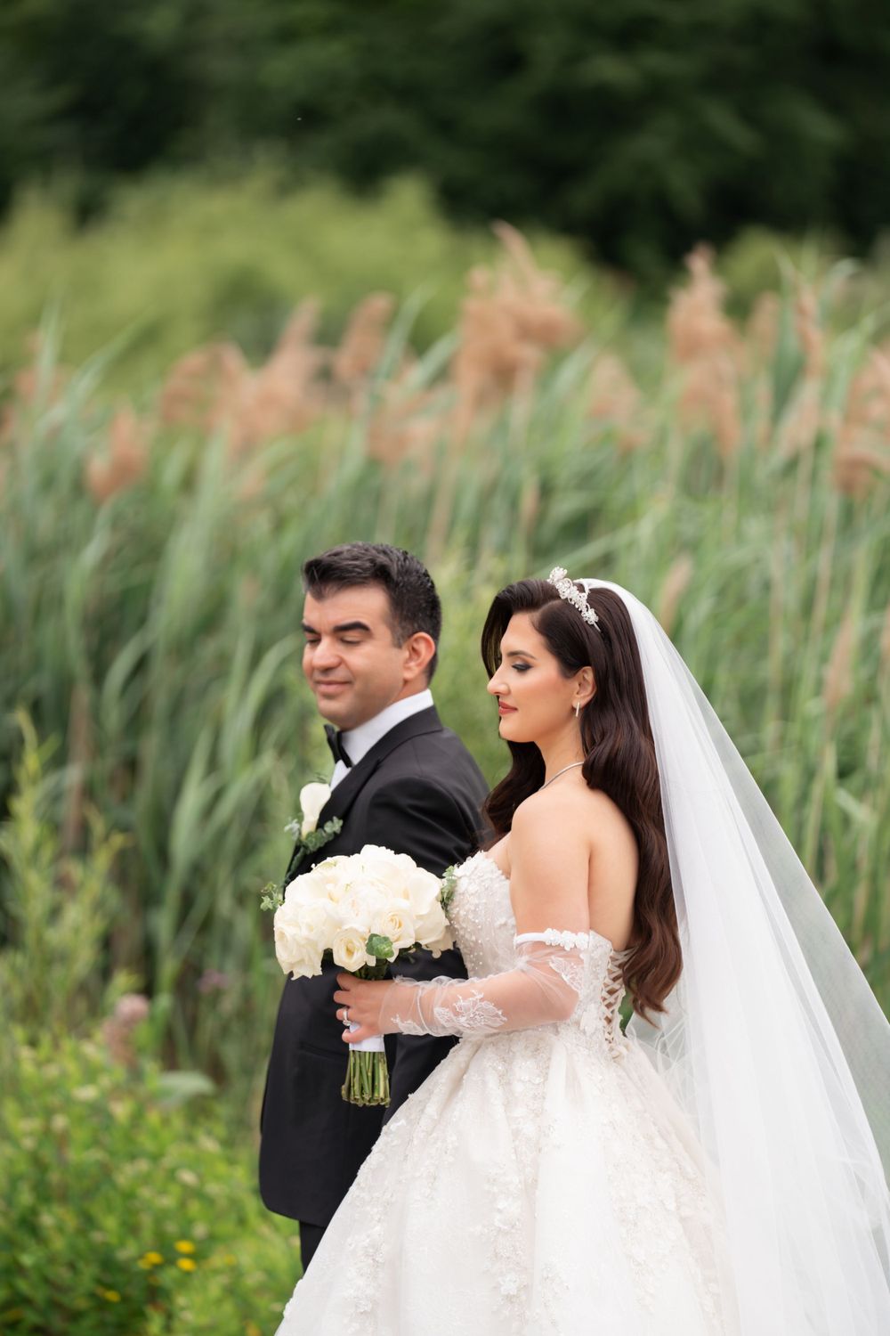 A bride in a white gown holds a bouquet while standing with her groom against a backdrop of tall reeds.
