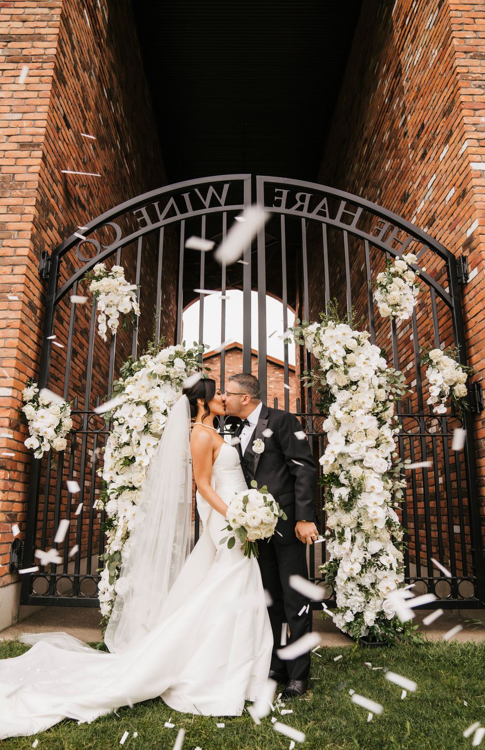 Wedding couple celebrating under floral arch gateway with white petals falling.