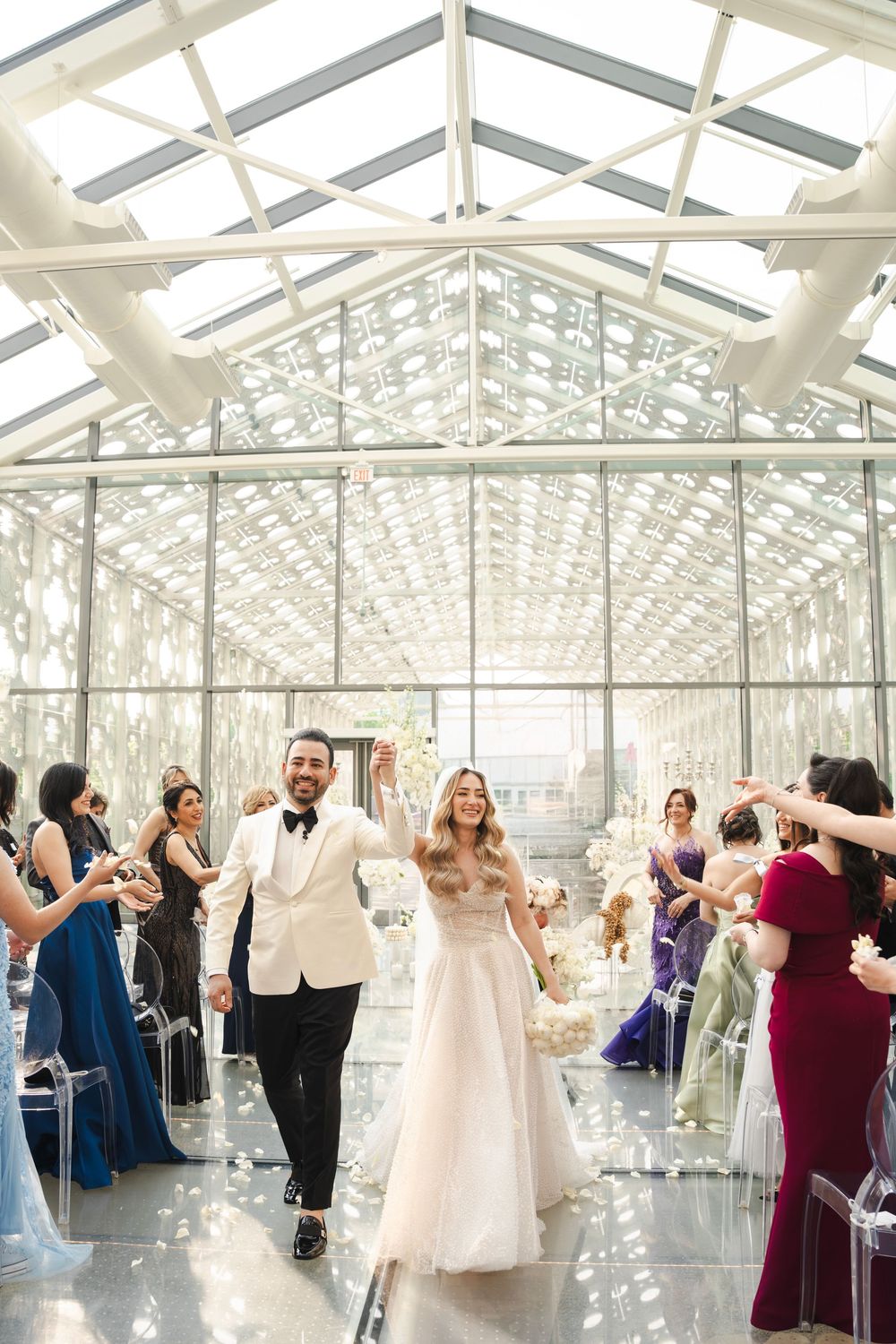 Wedding recessional in glass-ceiling venue with guests celebrating newlyweds walking down aisle.