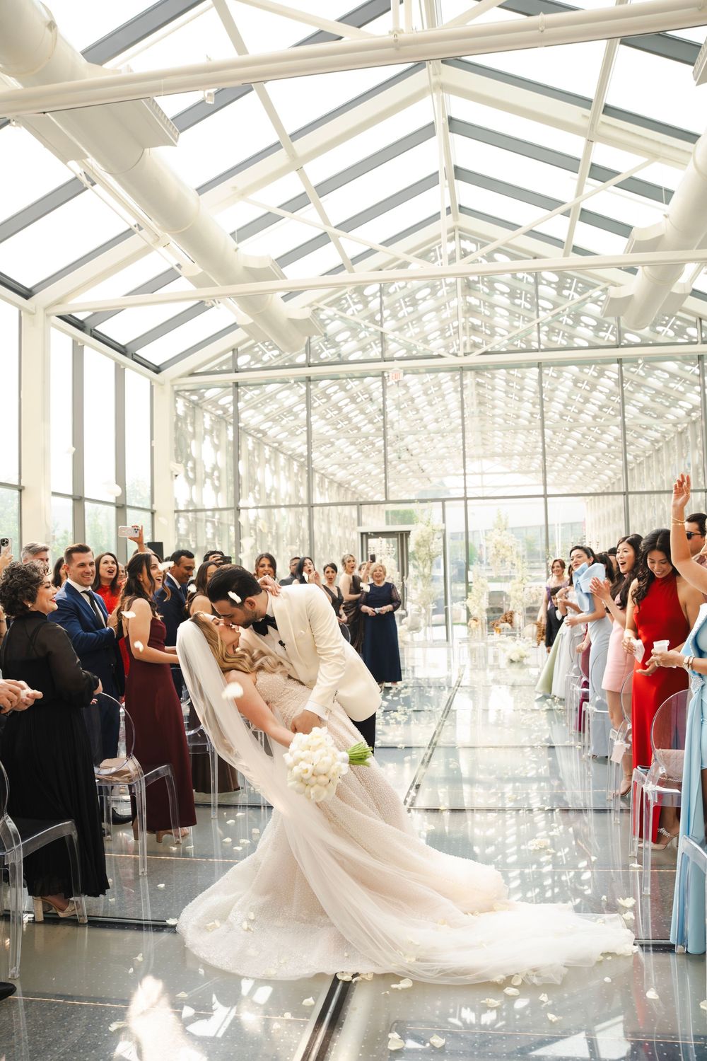 Wedding celebration in elegant glass-roofed venue filled with guests and flowing white decor.