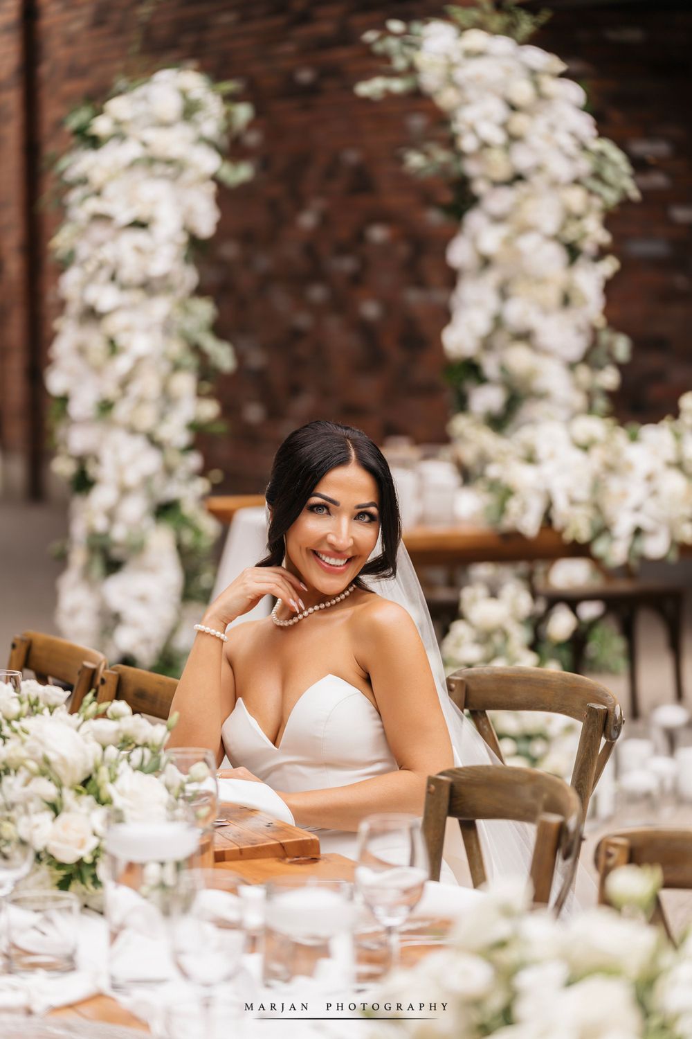 Bride seated at elegant outdoor reception table with white floral arrangements.