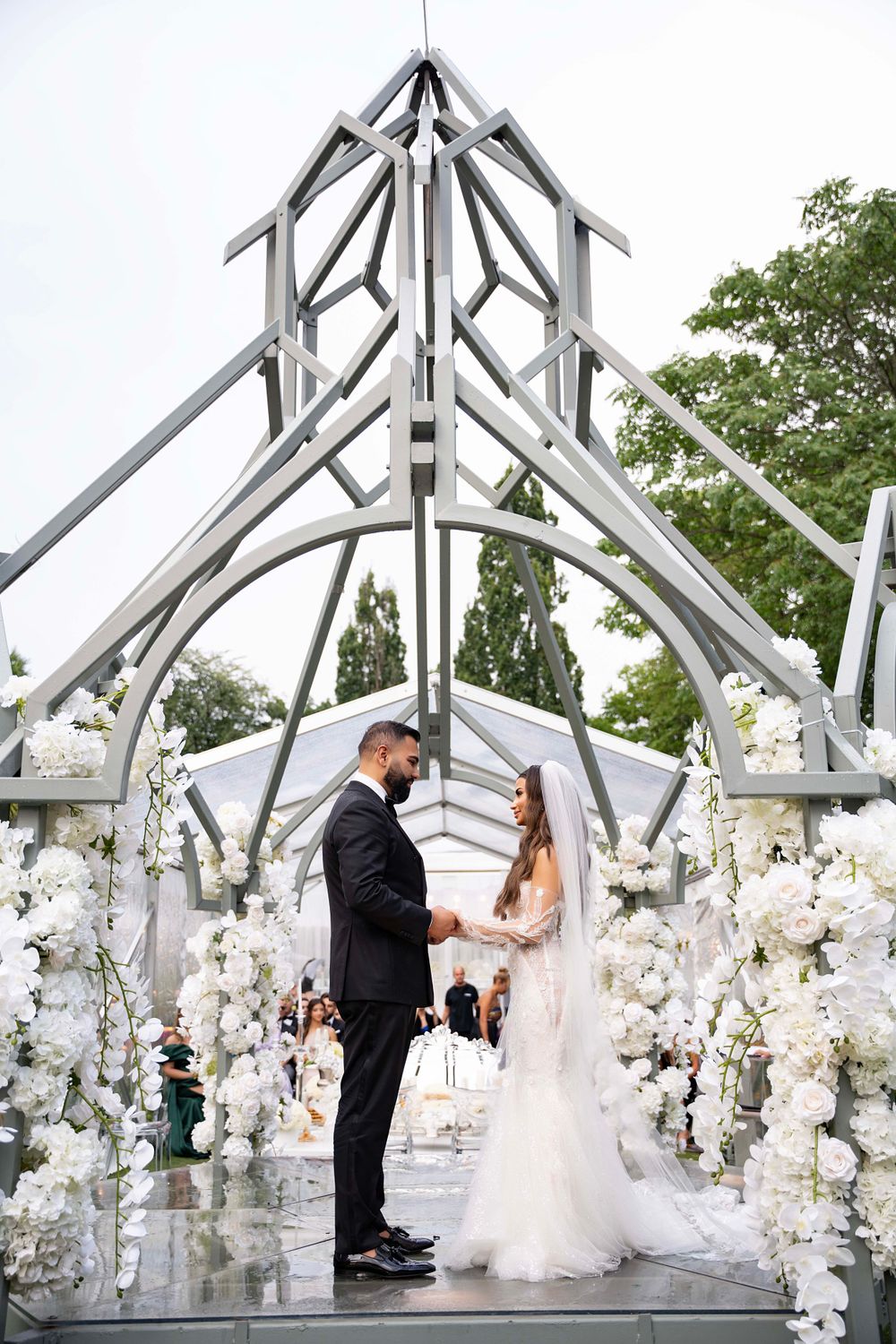 Modern wedding ceremony under geometric white arch decorated with white flowers.