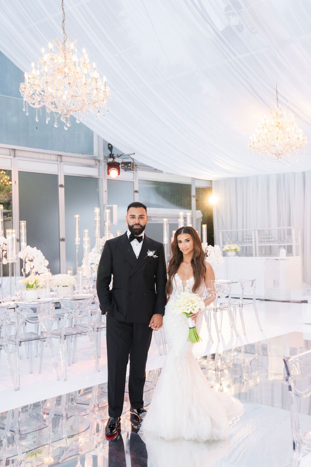 Modern white wedding reception venue with crystal chandeliers and ghost chairs.
