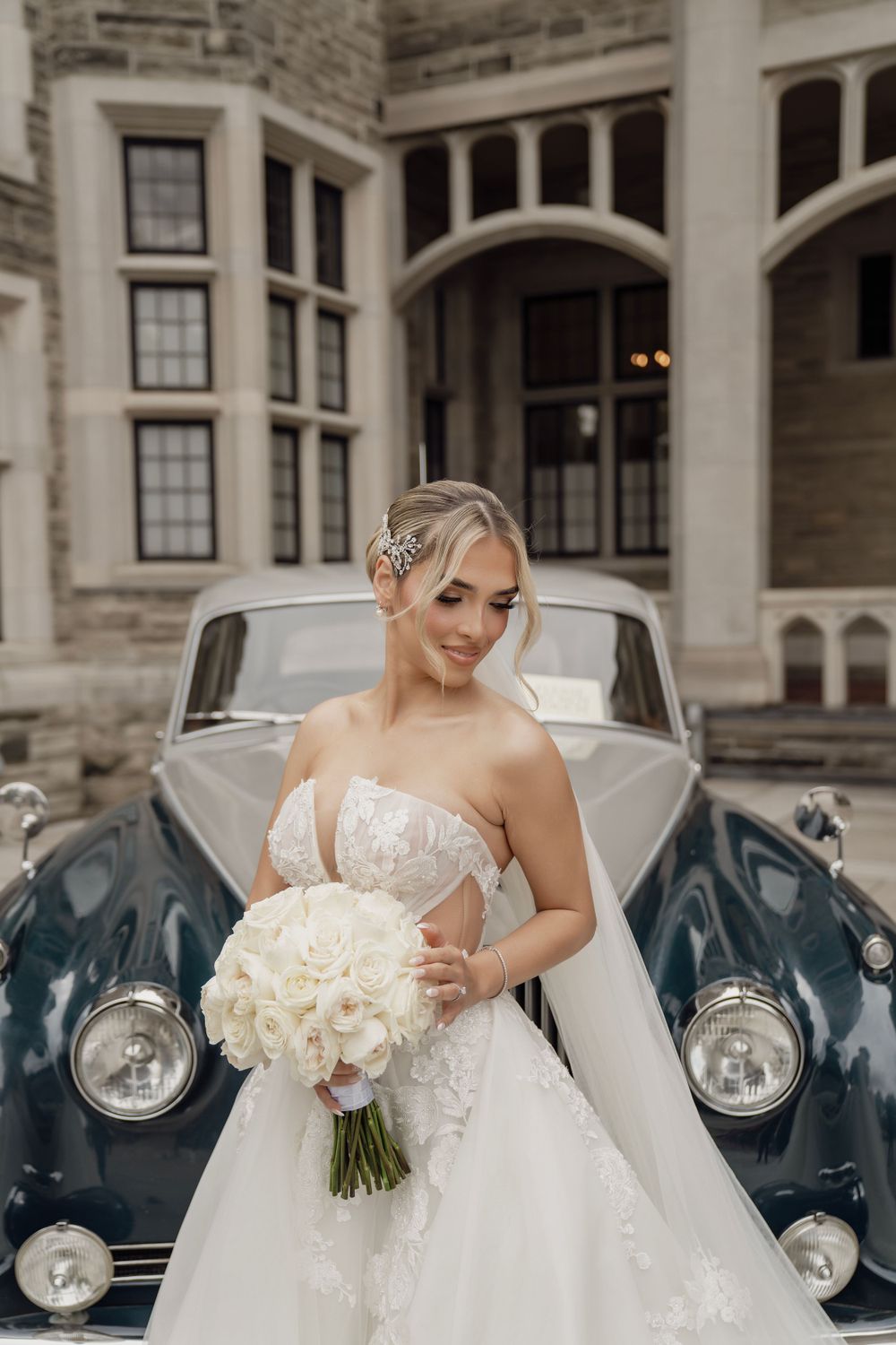 Elegant bride in white gown posing beside vintage blue car at stone architectural venue.