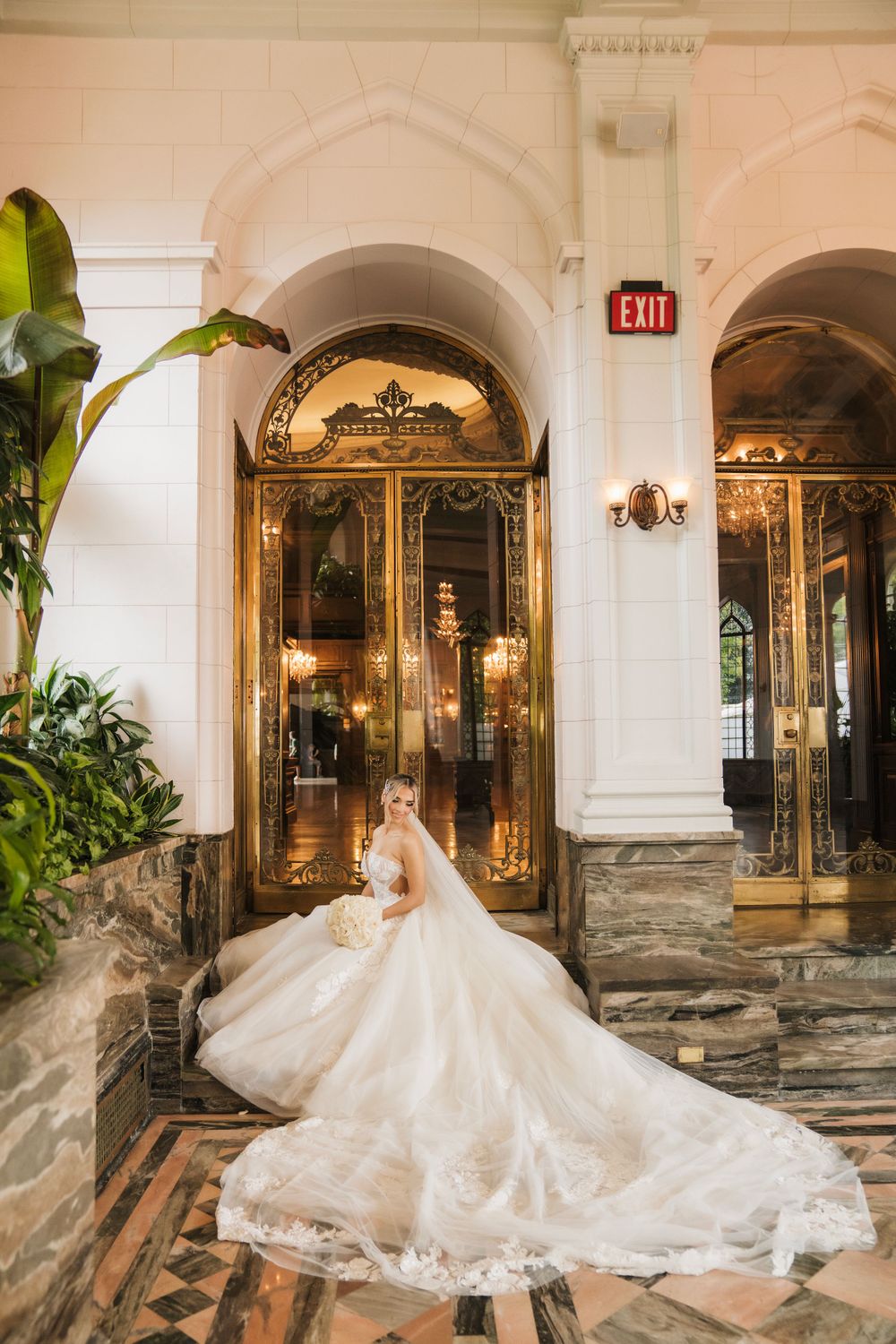Bride in flowing lace gown poses in opulent venue with ornate archways and mirrors.