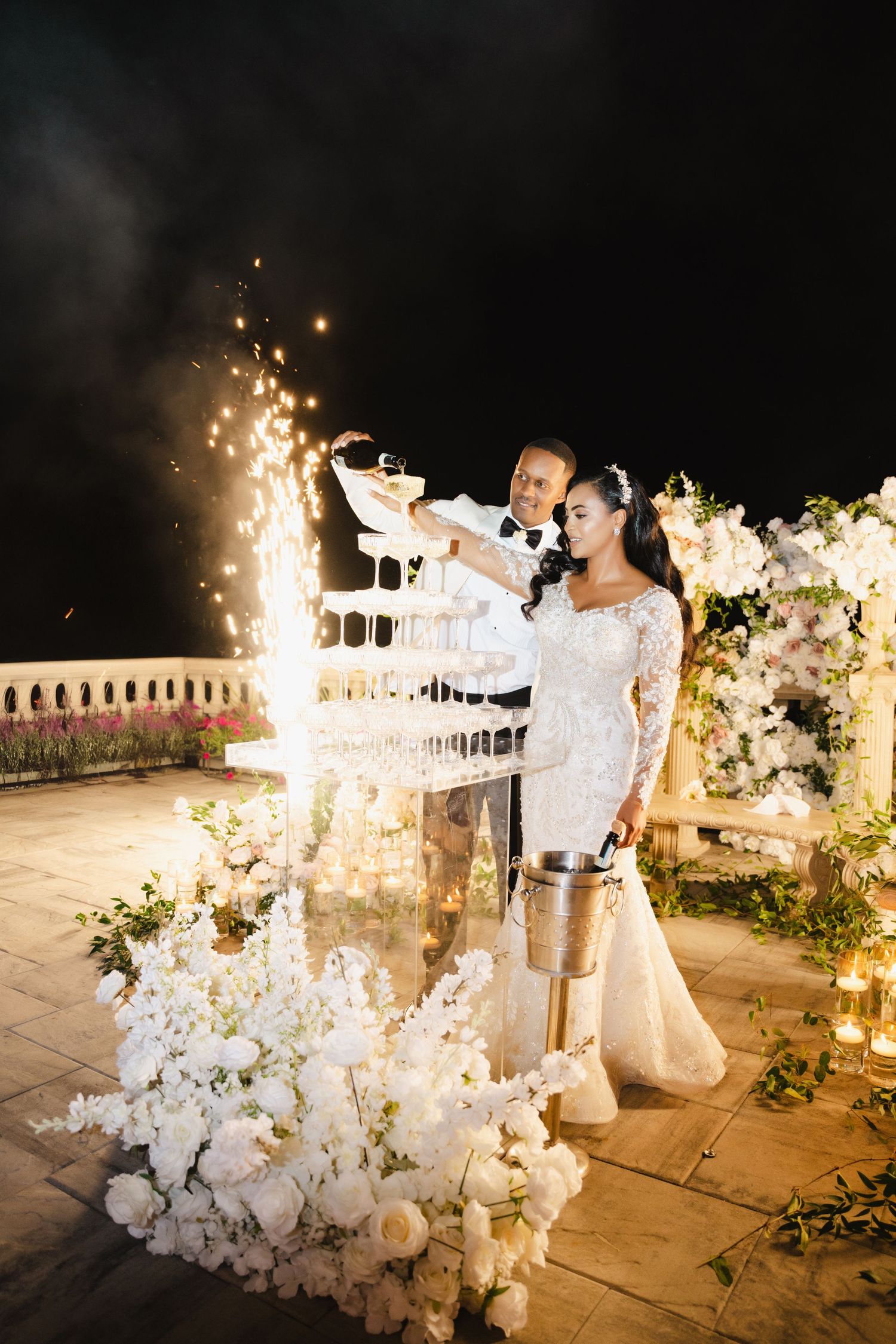 Couple celebrates with champagne tower and sparklers during nighttime wedding reception.