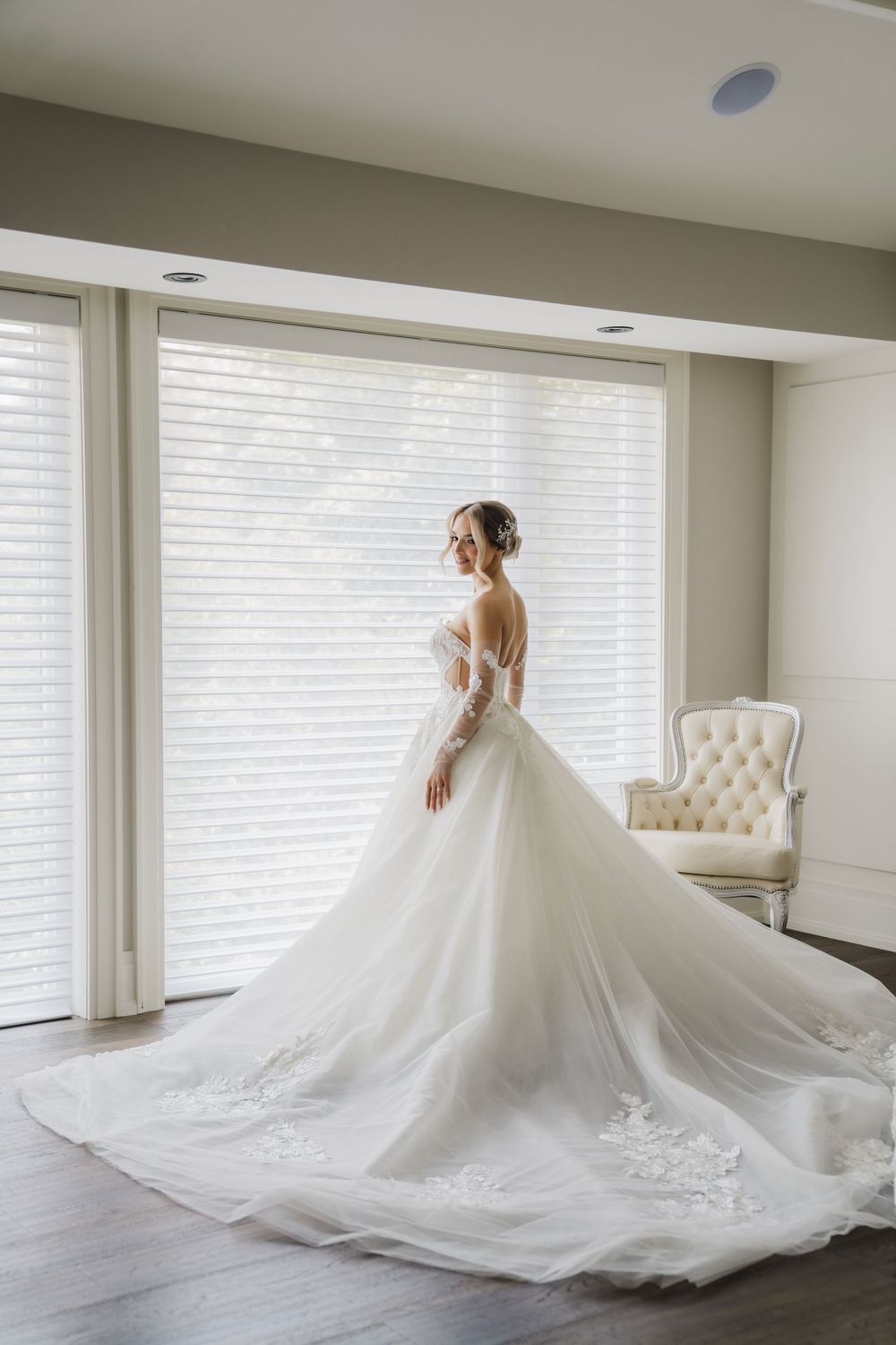 Bride in flowing white ball gown poses by large window in elegant room.