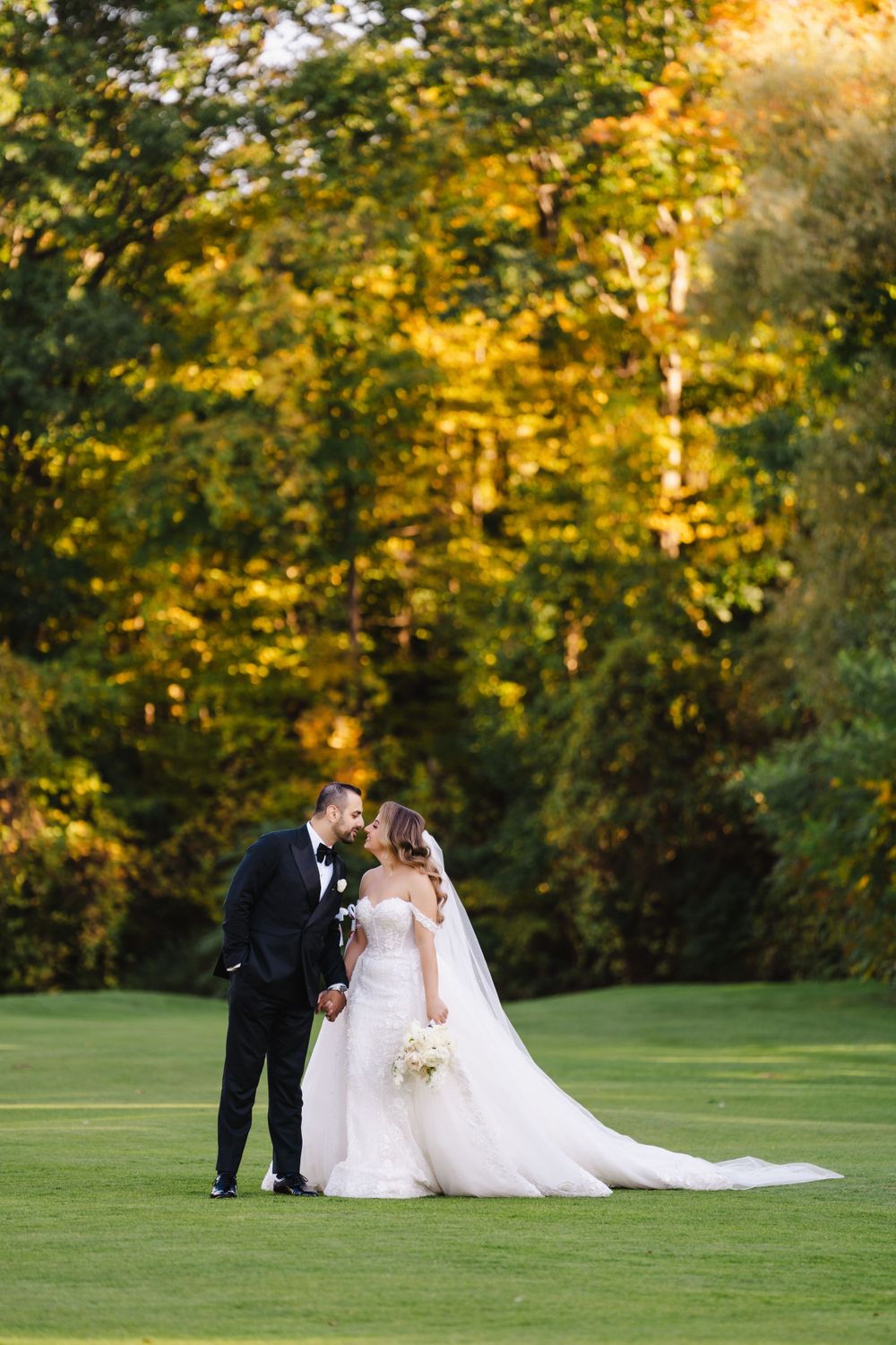 Newlywed couple poses on a golf course with golden autumn foliage in the background.