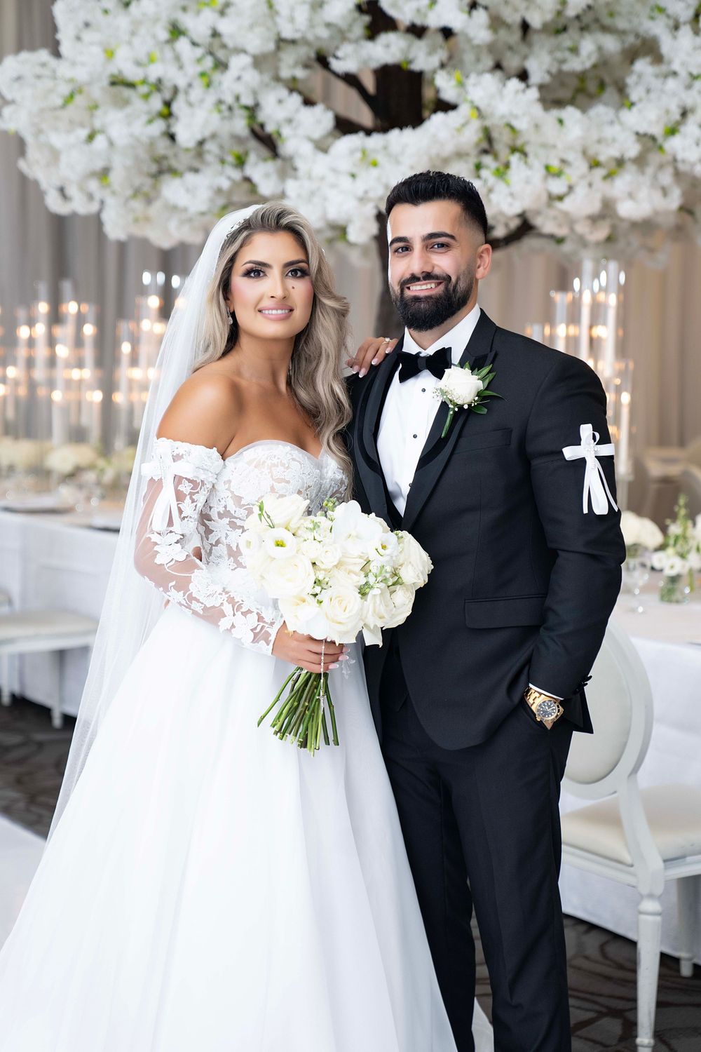 Elegant couple poses in formal wedding attire against white floral backdrop.