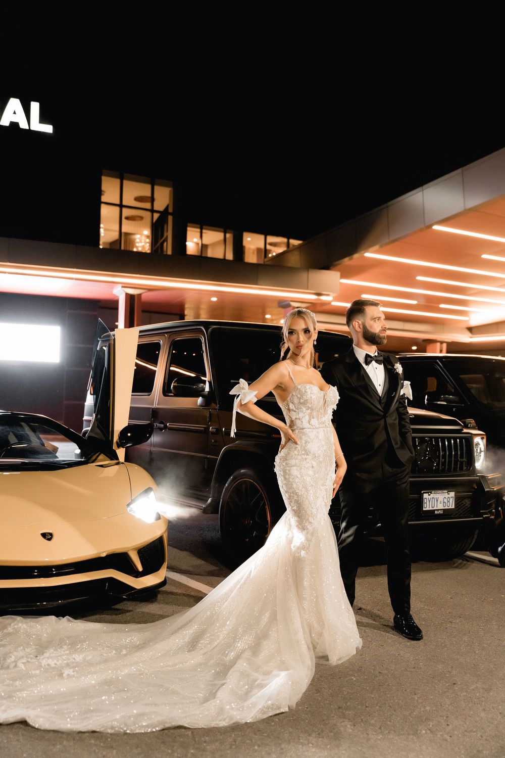 Nighttime wedding photo with bride's dramatic train trailing beside luxury vehicles.