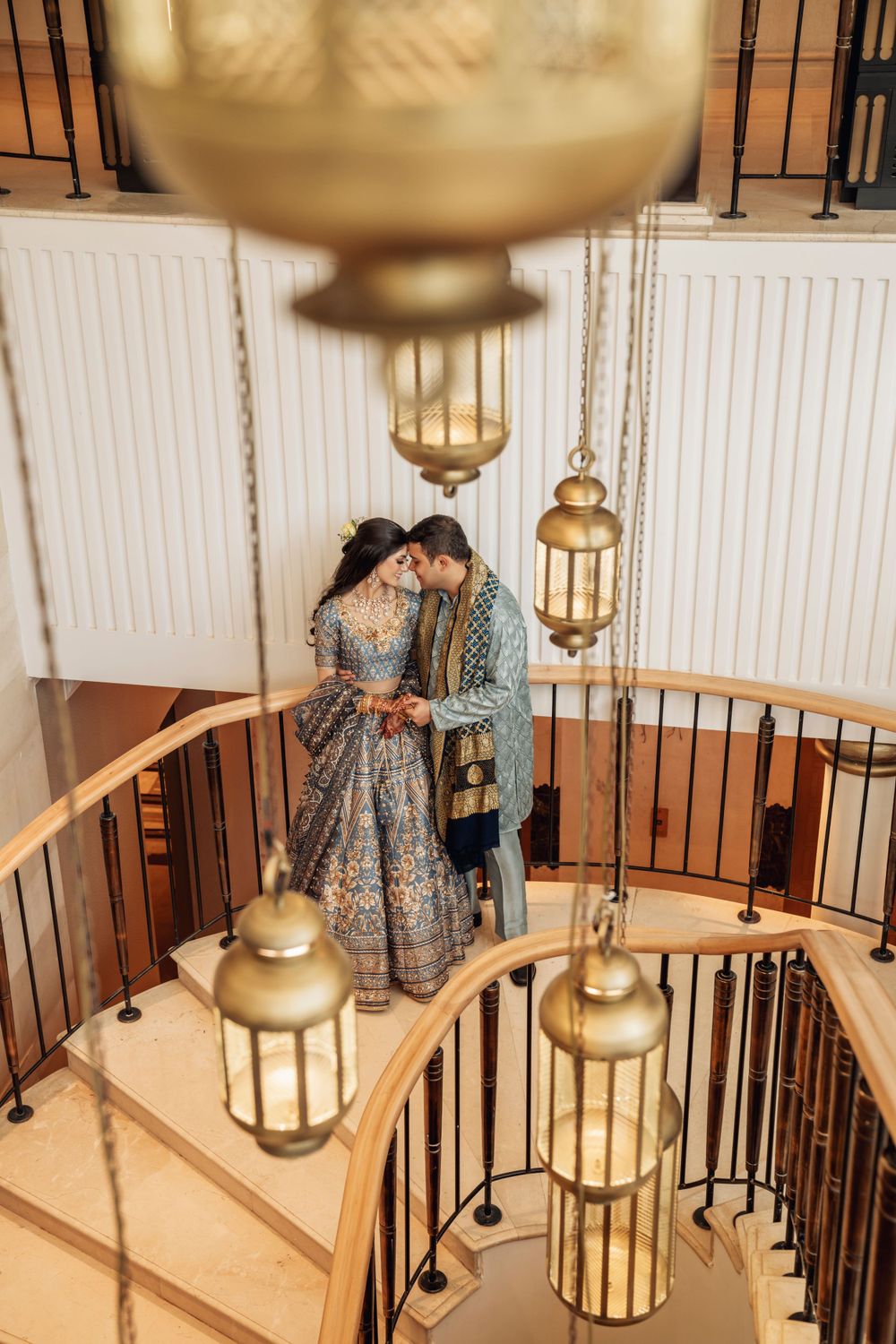 Couple in traditional Indian wedding attire embracing on spiral staircase with gold lanterns.