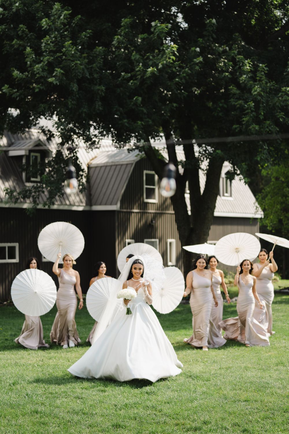 A wedding party poses with white parasols on a lush green lawn for a creative group photo.