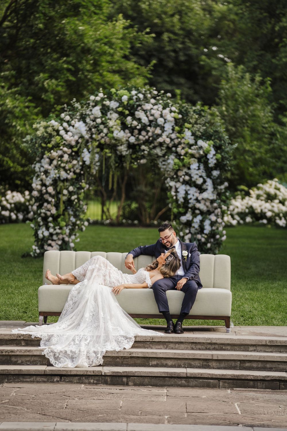 Newlyweds relax on white couch under floral arch in garden setting.