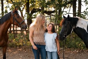 A mom and daughter stands together with their horses during golden hour with autumn trees in the background family photoshoot with horses