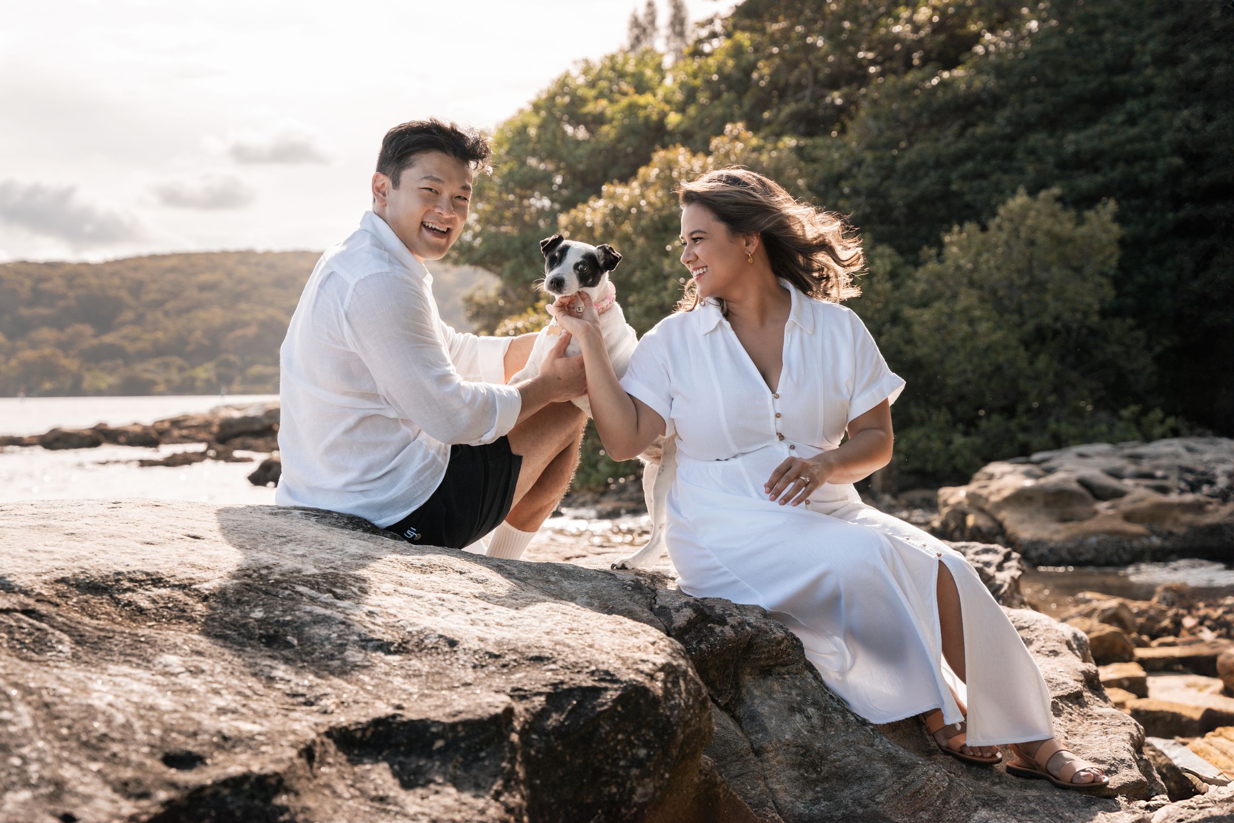 a man and a women sitting on a rock with their rescued cute dog in a nice backlit golden hour light during a photoshoot in Sydney