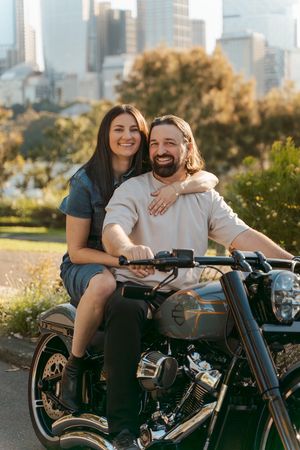 Couple sits together on a motorcycle in an urban setting with city skyline and trees in the background during proposal photoshoot in Sydney