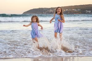 Joyful girls in purple dresses splash and play together in the ocean waves at sunset family photoshoot in Sydney Freshwater Northern Beaches