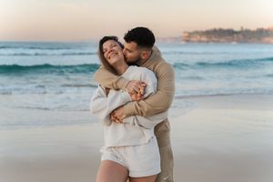 A couple shares a romantic moment on a sandy beach at sunset with ocean waves in the background Bondi Beach couples photoshoot